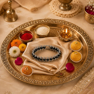 A silver-toned beaded bracelet with dark blue beads is displayed on a folded cream satin cloth inside a decorative gold-plated tray. The tray is styled with small sweets, flowers, a lit diya, and bowls of powder on a beige fabric surface.