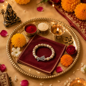 A beaded bracelet with a silver fish-shaped charm rests on a dark red cloth inside a decorative brass tray, surrounded by marigold flowers, a lit diya, small bowls of powder and rice, and a small statue on a warm beige surface.