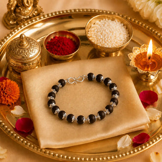 A black beaded rakhi with silver-toned spacer beads and a silver infinity clasp, displayed on a beige satin cloth over a decorative gold tray with bowls of powder, a lit diya, and flower petals.