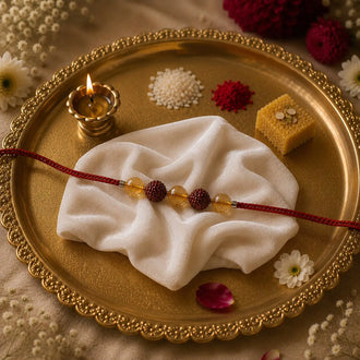 A red thread Rakhi with maroon and yellow beads is draped across a white cloth on a decorative gold tray, surrounded by a lit diya, small flowers, and sweets.