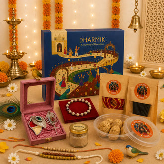 A festive Diwali gift set styled on a beige surface with warm string lights, marigold garlands, brass lamps, and a blue 'DHARMIK' box. The set includes a pink velvet box with silver jewelry, a beaded bracelet, packaged snacks, and a small jar, arranged decoratively with flowers and traditional props.