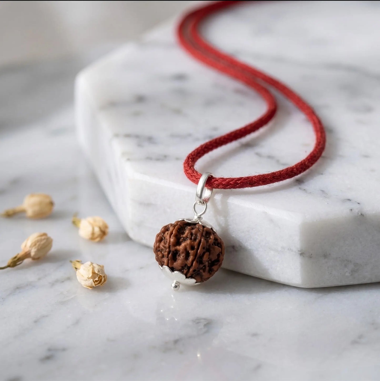 Silver-capped Rudraksha pendant on a red thread cord, displayed on a stone slab with fresh flowers