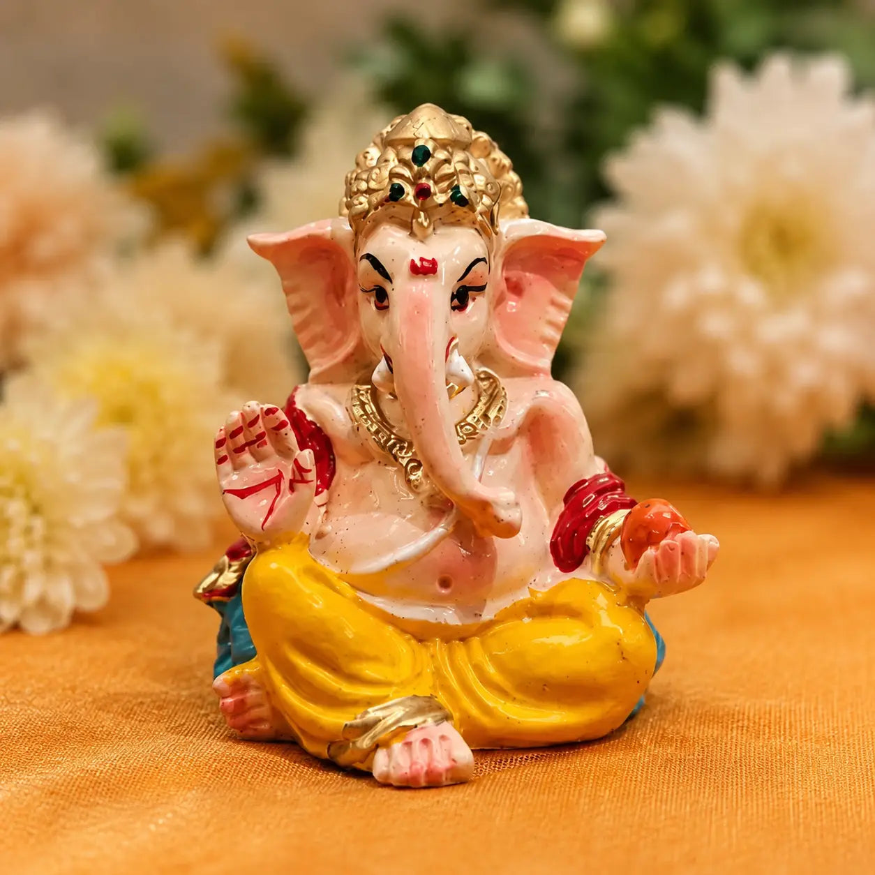 Hand-painted Lord Ganesha idol seated in a blessing posture, featuring a golden crown, yellow attire, and intricate details, placed on an orange cloth with a soft floral background.-  DharmikLife