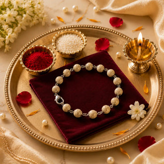 A beaded rakhi with cream-colored stone-like beads and silver-toned chain links is displayed on a dark red velvet cloth inside a gold tray. The tray is styled with small bowls of red powder and white beads, a lit brass diya, scattered rose petals, and cream fabric.