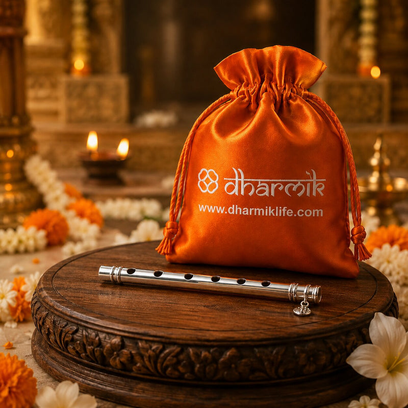 A silver metal flute rests on a carved wooden pedestal in front of an orange drawstring pouch printed with the Dharmik brand name and website. The background is a warm, softly blurred temple-like setting with lit candles and flowers.